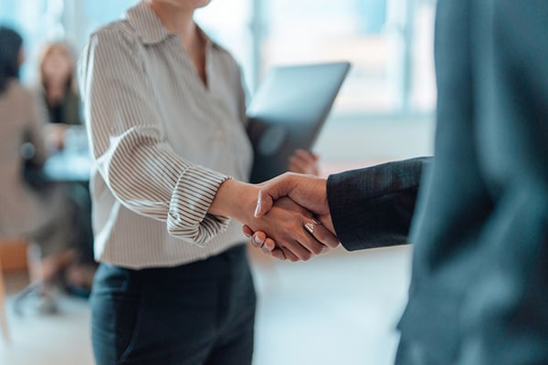 Two people shaking hands in an office holding a laptop