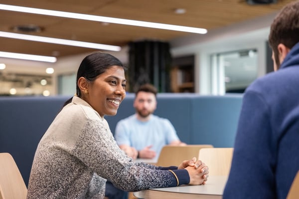 Two colleagues, a woman and a man, are sitting at a table, chatting in a comfortable office space.