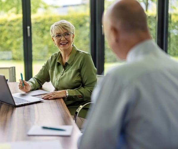 An older woman sat at a table with her laptop, smiling toward an older man