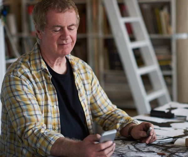 An older man in a chequered shirt, sat at a table, reading a text on him mobile phone