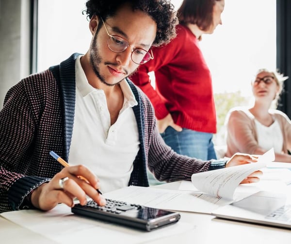 A close up of a man working using a calculator, while 2 women chat in the background