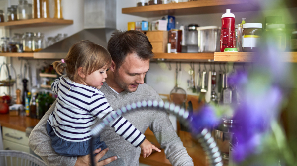 A man holds his young daughter while busy in the kitchen