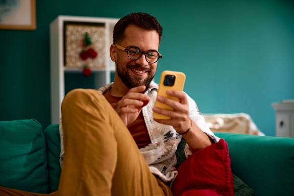 Young man using smartphone and smiling while relaxing on sofa at home