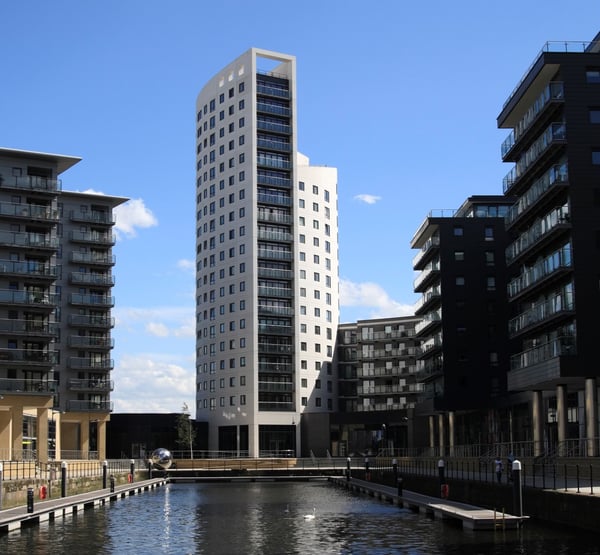 A tall grey building with lots of windows, next to smaller blocks of flats and a lake