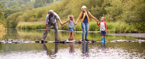 Grandparents with their 2 grandchildren, walking along a log on a river, holding hands