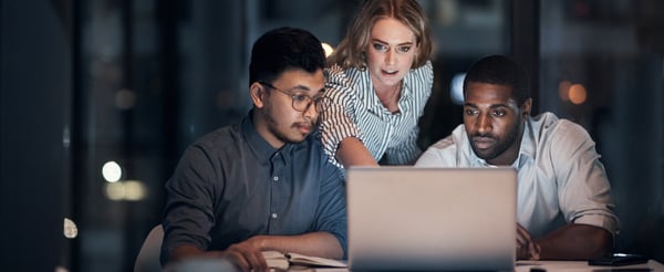 A woman and 2 men, working late at night in an office over a laptop