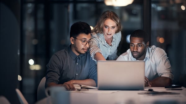 A woman and 2 men, working late at night in an office over a laptop