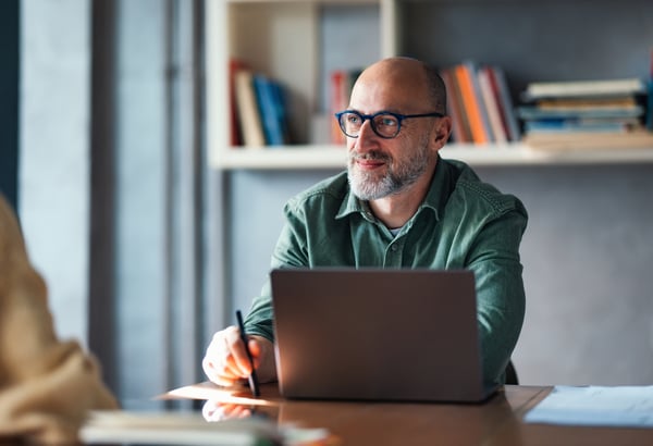 A older man with round glasses, sat at a desk with his laptop looking across the table