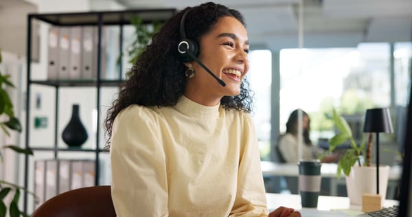 A young lady, in a pale yellow blouse working with a headset on and smiling