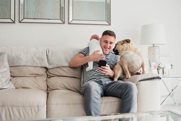 Man sitting on sofa holding baby and mobile phone with dog staring at him