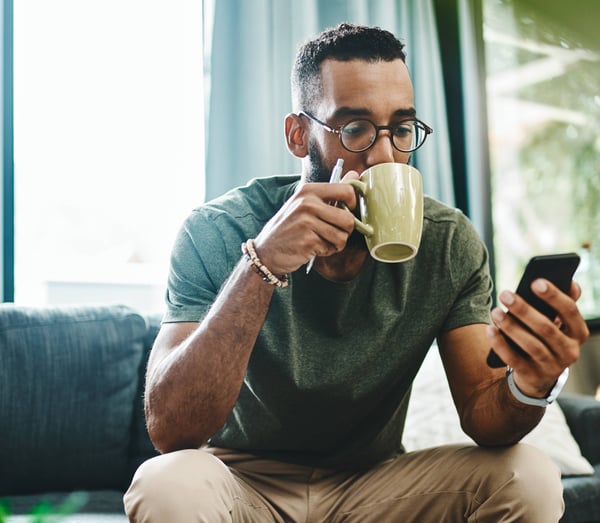 Man drinking coffee on sofa while looking at his mobile phone